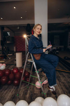 Smiling woman event planner in denim jumpsuit sitting on ladder writing notes while preparing festive balloons for event decorationの写真素材
