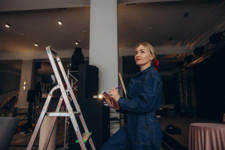 Woman standing on ladder at event venue, taking notes for setup and planning, orchestrating a successful occasionの写真素材