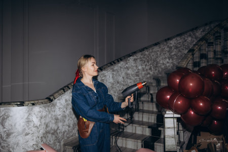 Young woman in denim jumpsuit melting balloons with a heat gun for event decoration on a marble staircaseの写真素材