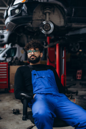 Male mechanic wearing blue overalls and safety glasses, sitting in a car repair shop with a car on a liftの写真素材