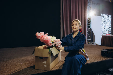 Woman florist in a denim jumpsuit preparing decorative flowers from a box, sitting on a stage in a backstage areaの写真素材