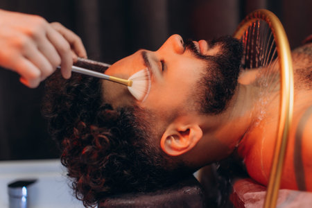 Young man relaxing during a wellness spa session, having a cosmetic product applied to his face with...
