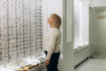 Senior woman standing in an optician shop, looking at a wall display of prescription spectacle...