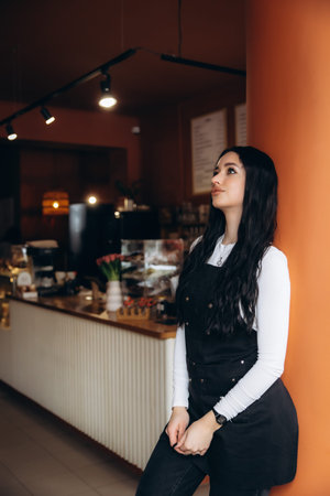 Young woman barista or waitress standing in a coffee shop, looking up, thinking about her small...