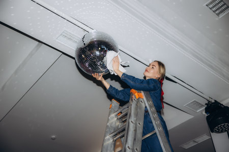 Young woman standing on an aluminum ladder, carefully wiping dust off a large silver disco ball hanging from the ceilingの写真素材