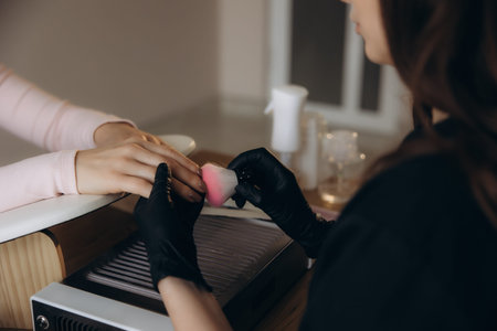 Manicurist wearing black gloves using a dust brush to remove residue from a client's hand, ensuring proper nail hygieneの写真素材