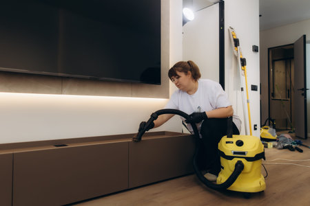 Professional cleaner wearing gloves vacuuming dust from television cabinet in modern apartment living room, ensuring thorough hygieneの写真素材
