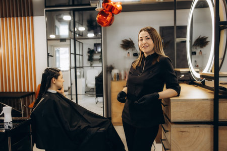 Smiling woman hairdresser standing in a modern beauty salon, welcoming a female client ready for professional hair care serviceの写真素材