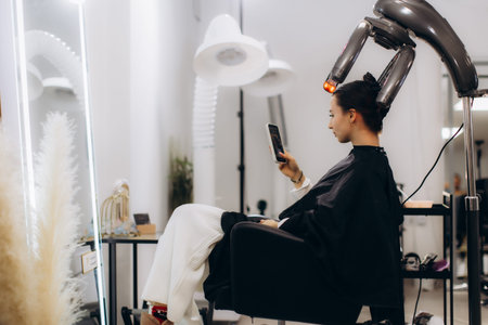 Woman in a modern hair salon having a hair treatment with a specialized infrared machine, using a smartphone while waiting