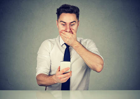 Casual young man covering mouth in shock while sitting and surfing smartphone realizing problem. の写真素材