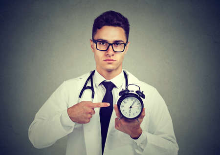 Young handsome man in medical uniform holding and pointing at clock looking confidently at camera. の写真素材