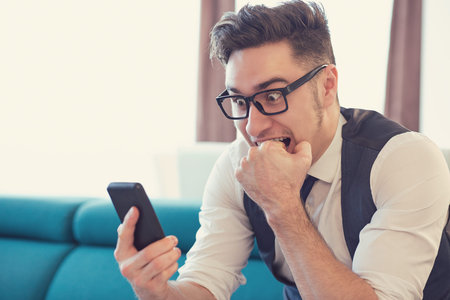 Young man in apartment watching smartphone and biting fist in expression of fear. 