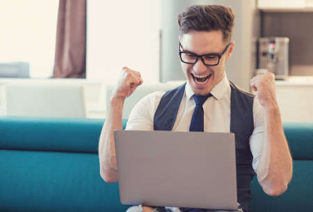 Young man using laptop and celebrating good news looking happy while sitting on couch. の写真素材