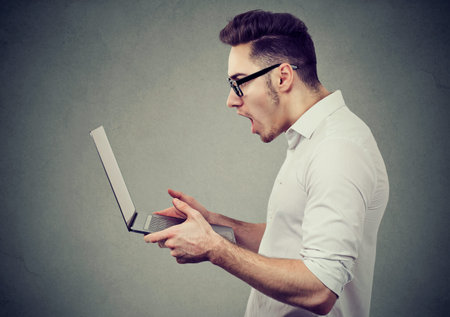Side view of man in eyeglasses holding and surfing laptop looking stunned while exploring latest...