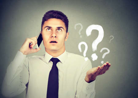 Misunderstanding and distant call concept. Upset annoyed young man talking on mobile phone has many questions isolated on gray background.の写真素材