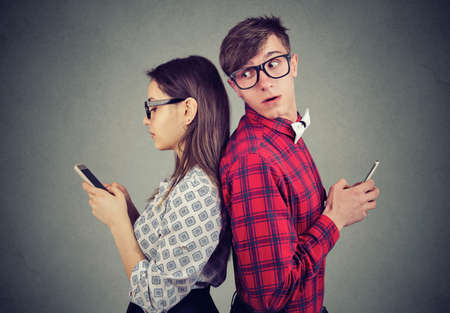 Young couple standing back to back, holding smartphones. Jealous man looking over his shoulder at his girlfriend phone trying to see what she is texting の写真素材