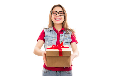 Charming young woman in glasses giving craft box with red bow and smiling at camera isolated on...