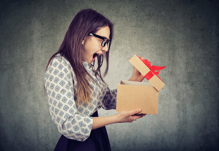 Side view of casual woman in glasses opening present box with bow looking super amazed on gray...