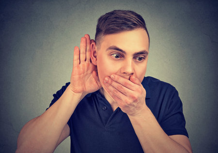 Young man holding hand near ear listening to gossips while covering mouth from amazement
