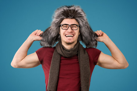 Young man wearing trendy furry winter hat and scarf looking happily at camera on gray background