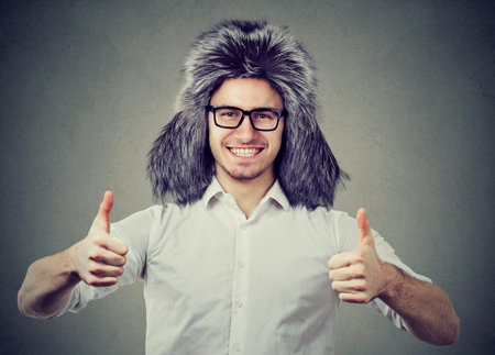 Young man wearing trendy furry winter hat and showing thumb up looking happily at camera on gray...