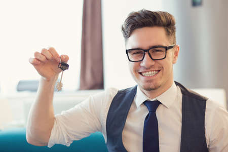 Young smiling man in eyeglasses holding key from new house sitting on couchの写真素材