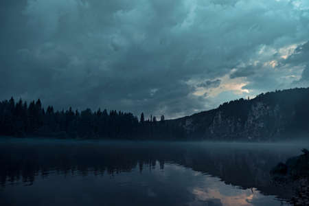 Night river on different banks of which there is a gloomy forest and rocks.の写真素材