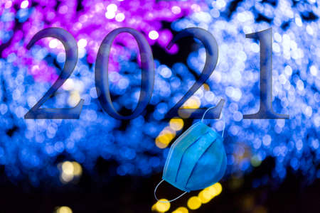 Text with the year number 2021 with a background of out-of-focus bright blue lights with Bokeh effect and a disposable blue hygienic face mask. Happy New Year 2021.の写真素材