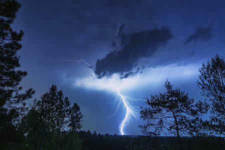 Lightning. Lightning strike. Spectacular night view of a lightning bolt streaking across the dark blue sky. Lightning and thunder and flashes. Clouds and silhouettes of treesの写真素材