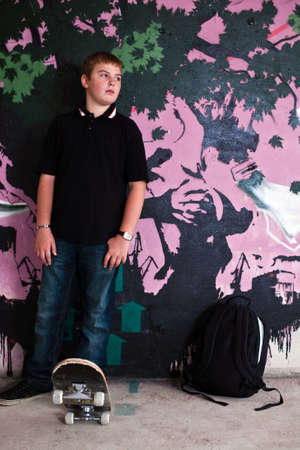 Young boy with shoulder bag and skateboard and graffiti wall in backgroundのeditorial素材