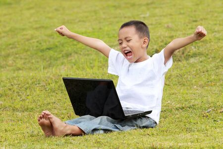happy young little boy celebrating success playing laptop on the grassの写真素材