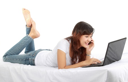 Happy girl on the phone lying in bed while using laptopの写真素材