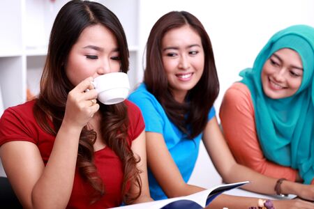 portrait of beautiful college student drinking a cup of tea and reading a book while her two friends looking at herの写真素材