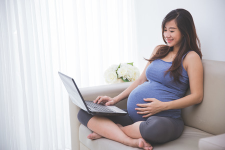 A portrait of a beautiful pregnant woman checking a while sitting on the couchの写真素材