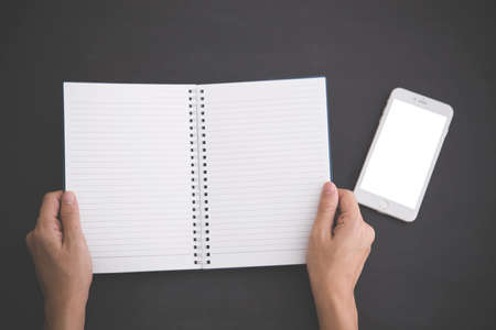 A portrait of hands open a notebook, and blank white mobile phone on the side. black background. Mock upの写真素材
