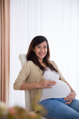 A portrait of a happy pregnant asian woman touching her big belly while sitting on a chairの写真素材