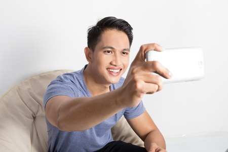 A portrait of a young attractive man taking pictures of him self (selfie) with smartphone while relaxing at homeの写真素材