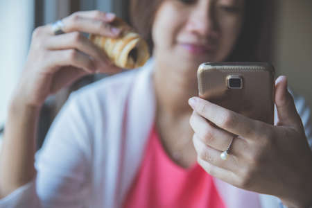 A portrait of Asian woman sitting hold a bread while using her handphone in cafeの写真素材