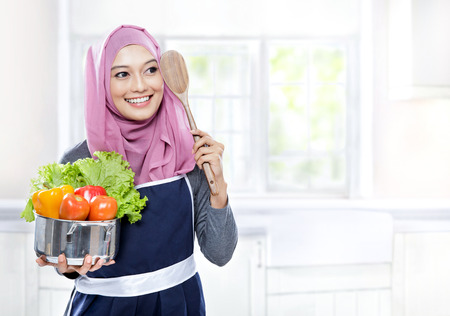 portrait of young woman carrying a pan full of vegetables and wooden spatula in the kitchen with copy spaceの写真素材