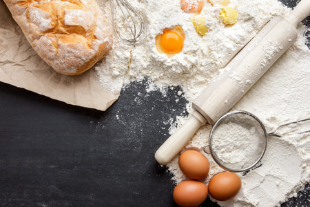 top view of rustic bread with egg, flour, steiner and rolling pin on black board with copy space