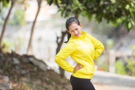 A portrait of a young asian woman exercising outdoor in yellow neon jacket, injured her waist