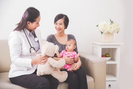 A portrait of an Asian female pediatrician showing a bear doll to the baby girl in mother lapの写真素材