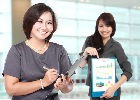 A portrait of a happy mature business women, with her assistant showing a graphic on clipboardの写真素材