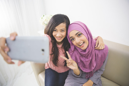 two happy young muslim woman take self portrait with handphone at homeの写真素材
