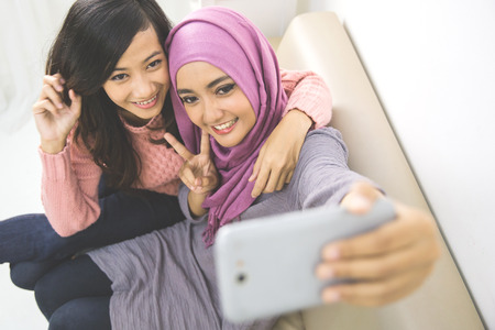 two happy young muslim woman take self portrait with handphone at homeの写真素材