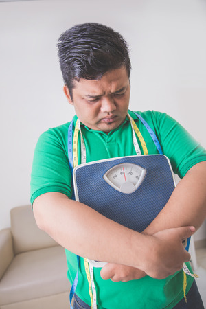 Portrait of sad obese man holding a weight scale, thinking about his weight problemの写真素材