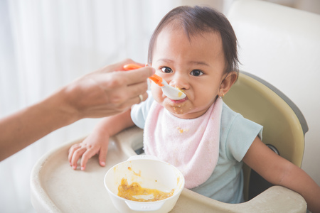 Portrait Of Happy Young Baby In High Chair being fed by mumの写真素材