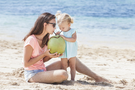 portrait of young mother sharing coconut water with her little daughter while sitting on the beach on summer vacationの写真素材