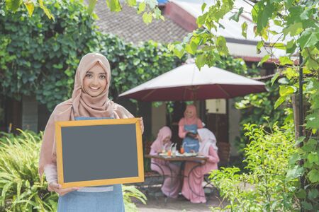 beautiful young asian waitress standing in front of a cafe and holding blank chalkboardの写真素材