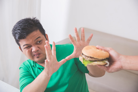 Portrait of a man close his mouth with hand refusing food while someone offering hamburger to himの写真素材